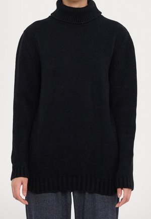Pull à col roulé côtelé noir porté sur un pantalon gris à motifs, visible des épaules aux hanches avec les mains détendues sur les côtés.