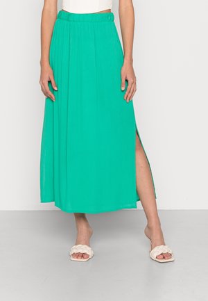 Kobieta ubrana w długą zieloną spódnicę z bocznym rozcięciem, biały top i białe, plecione sandały na otwartym palcu, stojąca na szarej podłodze na tle białej ściany.