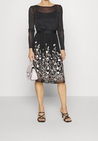 Frau in einem schwarzen, transparenten Langarmoberteil und einem schwarzen Rock mit floralem Stickerei, die eine hellrosa Handtasche hält und schwarze Absatzsandalen trägt.