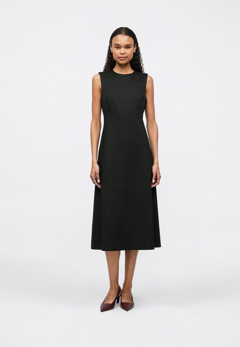 Žena stojící v bezramínkových černých midi šatech a burgundských lodičkách s špicí, na jednoduchém bílém pozadí.