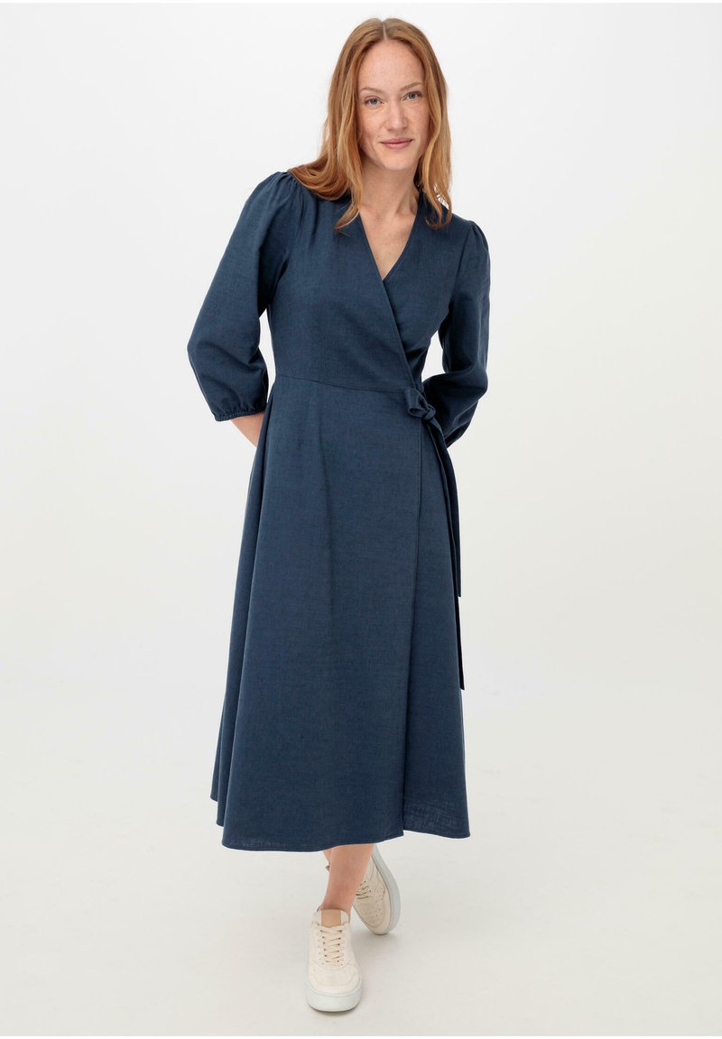 Dunkelblaues Wickelkleid mit Dreiviertelärmeln, tailliertem Gürtel und fließendem Rock; aus einem strukturierten Stoff; kombiniert mit weißen Sneakers.