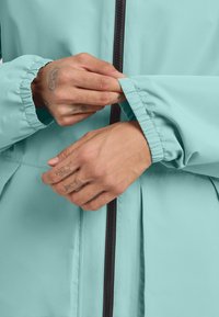 Veste bleu clair avec des poignets élastiques et une fermeture éclair noire. Le tissu semble lisse et légèrement brillant, avec une coupe décontractée et des détails de plis.