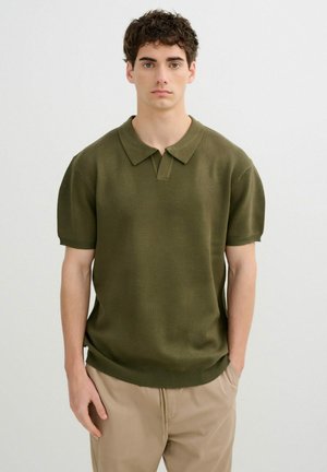 Joven con cabello rizado oscuro lleva una camisa polo de punto verde oliva de manga corta y pantalones beige, de pie frente a un fondo claro y liso.