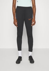 Pantalones de chándal negros con una cinturilla elástica, tobillos ajustados y un logo de Nike naranja en el muslo izquierdo. Llevados con zapatillas deportivas negras y calcetines blancos.