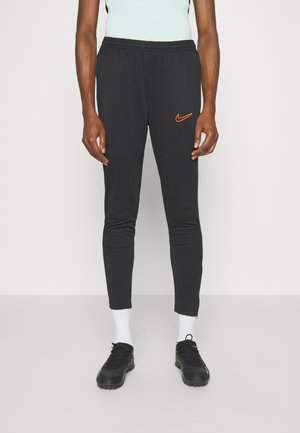 Czarne spodnie dresowe z elastycznym paskiem w talii, dopasowanymi kostkami i pomarańczowym logo Nike na lewej udzie. Nosi się je z czarnymi sneakersami i białymi skarpetkami.