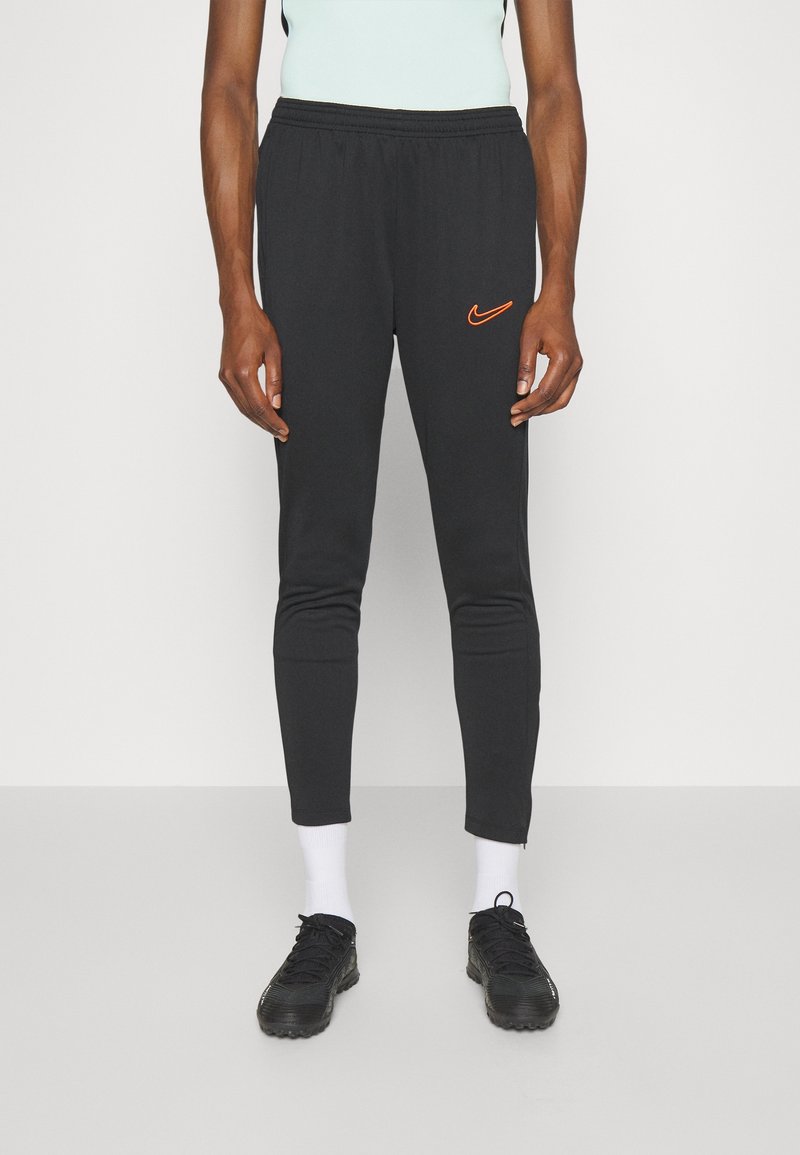 Pantalones de chándal negros con una cinturilla elástica, tobillos ajustados y un logo de Nike naranja en el muslo izquierdo. Llevados con zapatillas deportivas negras y calcetines blancos.