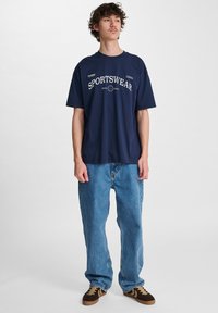 Marinblå bomullströja med "SPORTSWEAR" grafik, i kombination med löst sittande ljusblå jeans och bruna sneakers med svarta och gula detaljer.