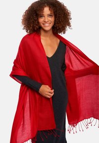 Châle rouge en tissu léger, avec un ourlet à franges, drapé sur une robe noire à col en V, mettant en valeur une texture lisse.