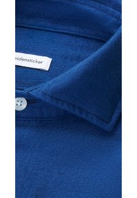 Colletto di camicia in cotone blu con una superficie liscia e textured. Presenta un'etichetta bianca e una leggera chiusura con bottone sul colletto.