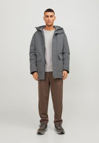 Grå polstret jakke med hætte, to frontlommer og lynlås, lagdelt over en lysegrå sweater og brune bukser.