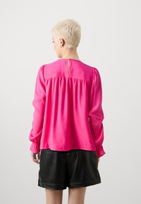 Blusa fucsia con maniche lunghe, design con schiena arricciata e dettaglio a chiave al collo, abbinata a pantaloni corti in finta pelle nera.