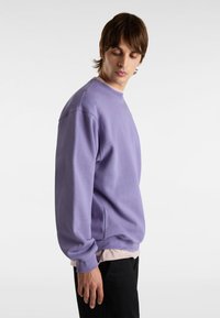 Sudadera de color púrpura claro con cuello redondo y corte oversized, con mangas largas y puños acanalados, llevada sobre una camisa clara.
