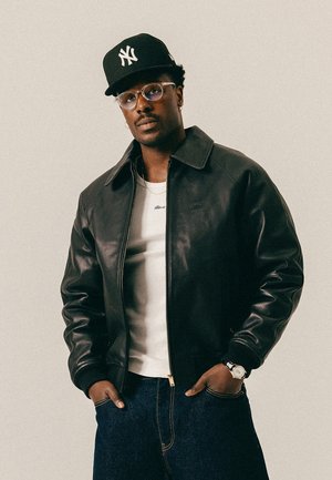 Mann trägt schwarze New York Yankees Kappe, klare Brille, schwarze Lederjacke, weißes Hemd, dunkle Jeans, Uhr und Creolen, Hände in den Taschen.