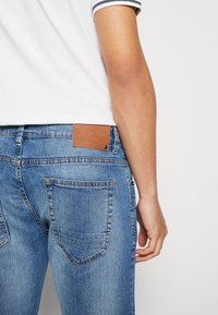 Jeans de mezclilla azul con un parche de cuero en la parte trasera etiquetado como "INDIE DUE", que presenta dos bolsillos traseros y un acabado ligeramente descolorido.