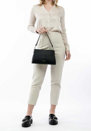 Vrouw die een beige blouse en broek draagt, met een zwarte leren handtas met korte handvatten, gecombineerd met zwarte loafers.