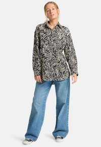 Camisa de botones hecha de una tela con un patrón abstracto en blanco y negro. Mangas largas, diseño con cuello, combinada con unos vaqueros de pierna ancha en azul claro.