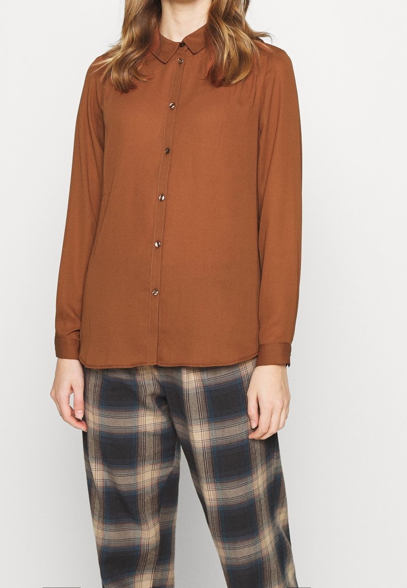 Chemise marron à manches longues avec un col et une fermeture à boutons. Associée à un pantalon à carreaux dans des tons beige, bleu et marron. Texture de tissu lisse.