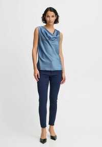 Mouwloze top met kraag in lichtblauwe satijn, gecombineerd met nauwsluitende marineblauwe broek en zwarte pumps met spitse neuzen. Gladde textuur en elegante val.