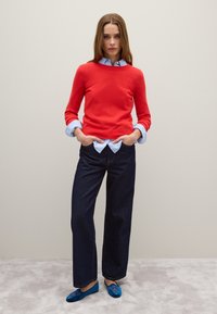 Maglione rosso, camicia azzurra con colletto sotto, jeans larghi blu scuro, ballerine blu con ricami floreali, in piedi su un tappeto grigio chiaro.
