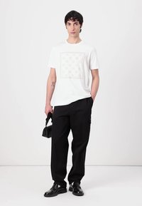 Camiseta blanca con un gráfico sutil en el frente, combinada con pantalones anchos negros y zapatos negros, sosteniendo un bolso negro.