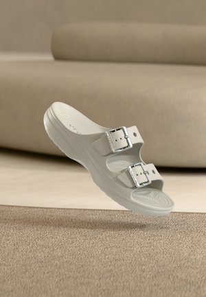 Ανοιχτό μπεζ σανδάλι slip-on με δύο ρυθμιζόμενες ασημένιες αγκράφες, υφασμένο πέλμα και ανυψωμένη σόλα, να αιωρείται πάνω από μια υφασμένη μπεζ μοκέτα.