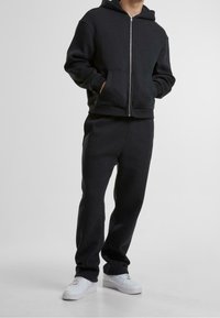 Persona che indossa una felpa nera con zip e pantaloni tuta neri abbinati, con sneakers bianche, in piedi su uno sfondo chiaro uniforme.