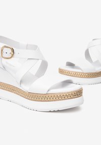 Hvide wedge-sandaler har et stropdesign, gylden fletdetalje rundt om sålen og en glat tekstur. Justerbar spænde lukning er inkluderet.