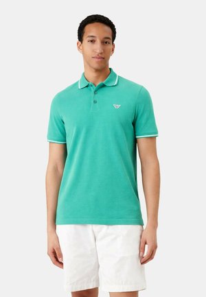 Mężczyzna stoi, ubrany w turkusową koszulkę polo z krótkim rękawem i białym obszyciem oraz białe szorty na jednolitym tle.