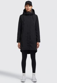 Svart lång parka med huva, justerbara dragsnören, fickor framtill och nedre kant som är kortare fram än bak, kombinerad med svarta leggings och vita sneakers.