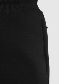 Schwarze Sweatpants aus weichem Stoff, mit einem gerippten Bund und einer Seitentasche mit einem kleinen metallischen Akzent. Glatte Textur.