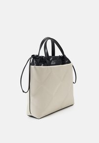 Sac fourre-tout matelassé beige avec des poignées doubles en cuir noir et une pochette à cordon noire à l'intérieur, sur fond blanc.