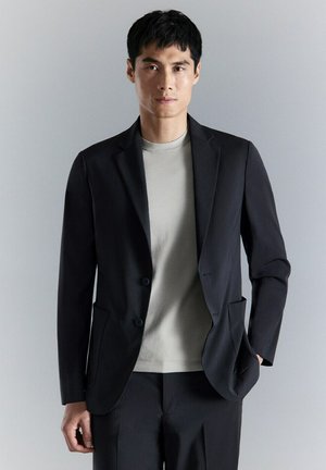 Uomo che indossa giacca e pantaloni neri con maglietta girocollo grigio chiaro, in piedi davanti a uno sfondo grigio chiaro semplice.