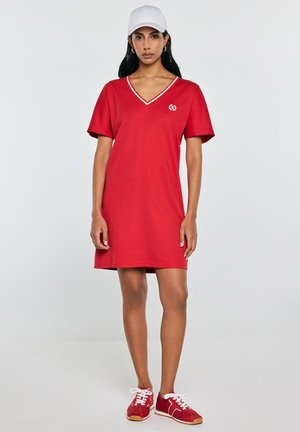 Kobieta ubrana w czerwoną sukienkę z dekoltem w serek i krótkim rękawem, białą czapkę z daszkiem i czerwone trampki, stojąca na jednolitym tle.