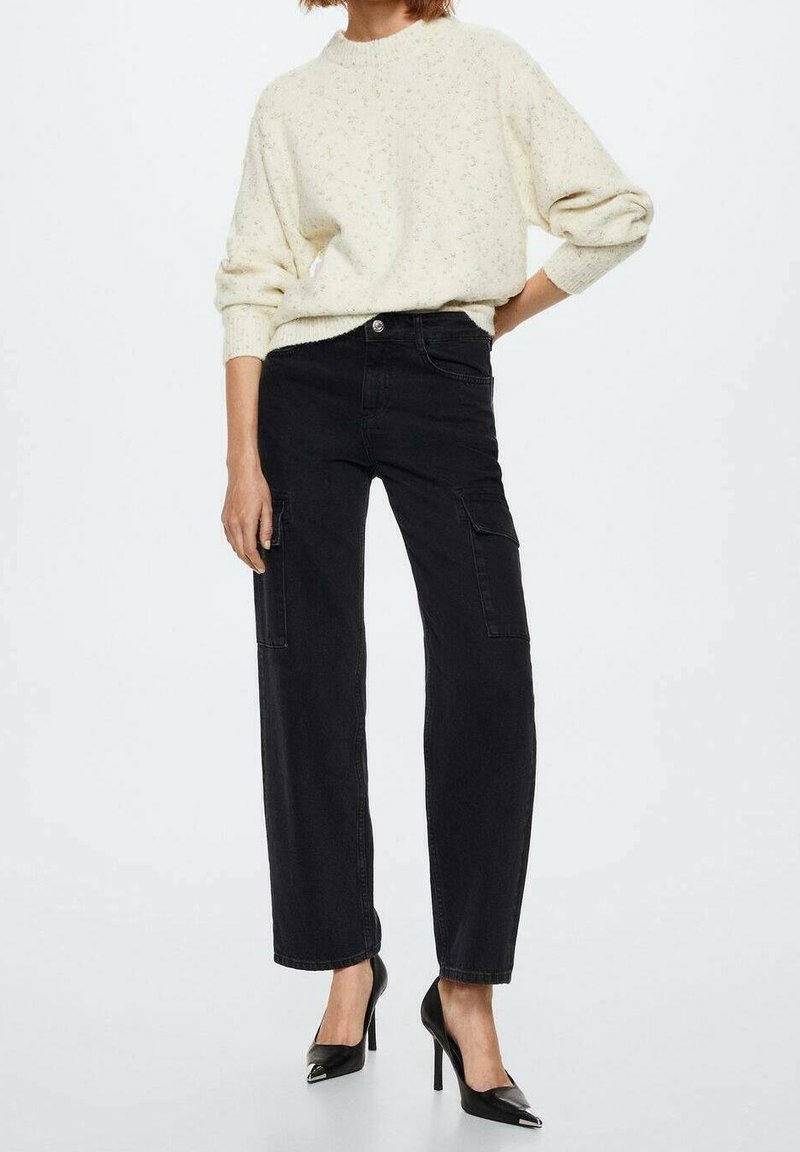 Femme portant un pull en maille crème, un pantalon cargo noir taille haute et des escarpins noirs à bout pointu, debout avec une main sur la hanche.