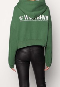 Persona che indossa un cropped hoodie verde con testo bianco sul retro e pantaloni di pelle nera aderenti, in piedi contro uno sfondo neutro.