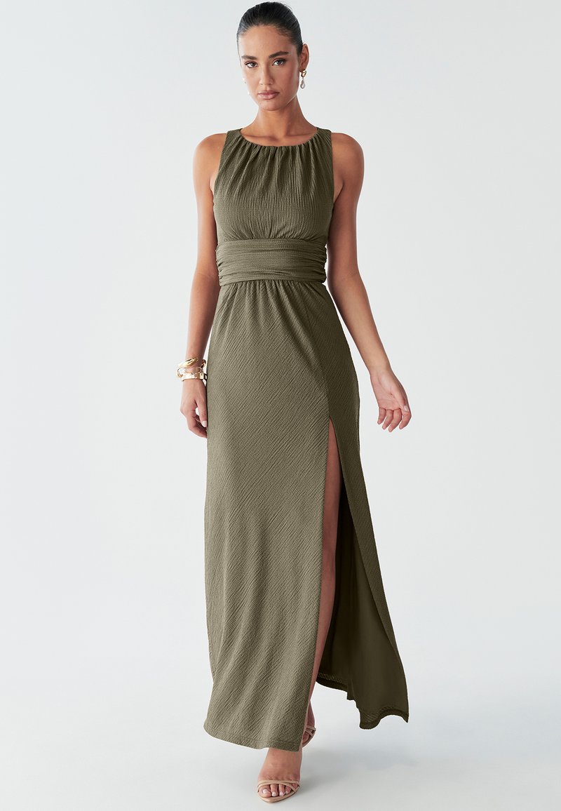 Model in einem langen olivgrünen Kleid mit hohem Schlitz, die selbstbewusst mit einem leichten Lächeln posiert und passende Schmuckstücke sowie Sandalen trägt.