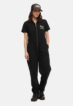 Femme portant une combinaison noire zippée avec un logo tigre et une casquette Von Dutch noire et imprimé léopard assortie, debout les mains dans les poches.