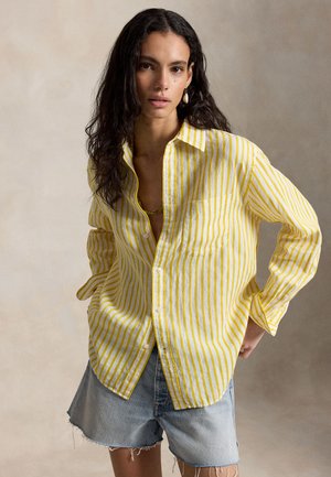 Γυναίκα με μακριά κατσαρά μαλλιά, φορώντας πουκάμισο με κίτρινες και λευκές ριγέ λεπτομέρειες και ανοιχτόχρωμα τζιν σορτς, στέκεται με το ένα χέρι πίσω από την πλάτη της.