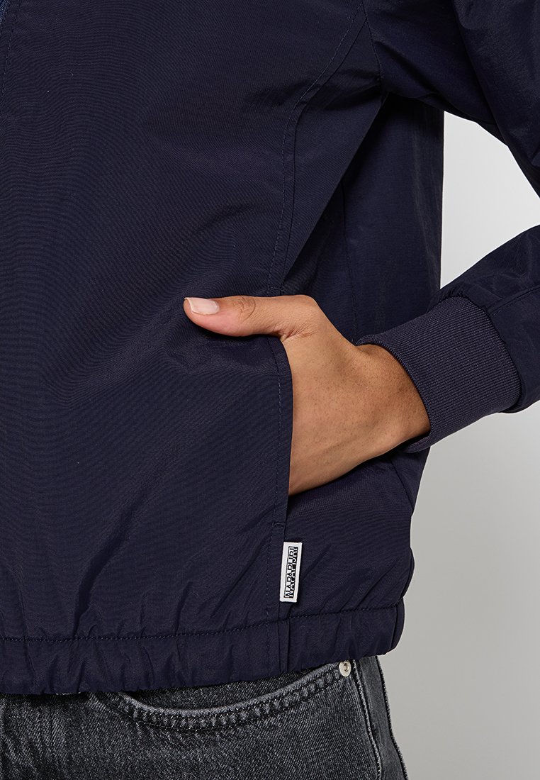 Giacca a vento navy con una texture liscia e leggera, polsini elasticizzati, tasche laterali e una piccola etichetta del marchio all'orlo.