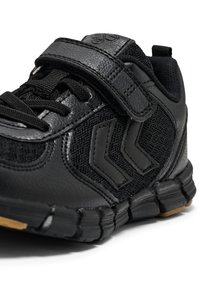 Zwarte sportieve schoen met een gestructureerde mesh bovenkant, accenten van synthetisch leer, klittenbandsluiting en rubberen zool. Heeft een gebogen ontwerp en een chevronlogo.