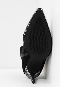 Musta mokkanahkainen korkokenkä, jossa on terävä kärki, koristeltu hopeisilla niiteillä kantapään ympärillä ja tekstuurilla varustettu kumipohja.