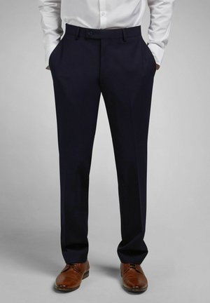 Mann trägt marineblaue Anzughose, weißes, eingestecktes Hemd, braune Lederschuhe, Hände in den Taschen, steht vor schlichtem grauem Hintergrund.