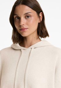 Jeune femme avec les cheveux bruns courts portant un sweat à capuche beige sur un fond blanc uni.