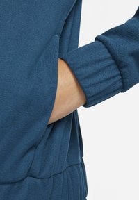 Blå pullover med blød tekstur, ribbestrikkede manchetter og sidelomme. Stoffet virker let og let fluffy.