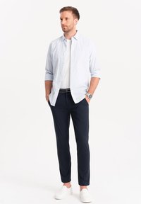 Camicia button-up a righe blu chiaro sopra una maglietta bianca, abbinata a pantaloni scuri e scarpe bianche senza lacci. Orologio d'argento al polso.