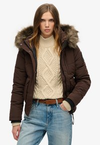 Bruine pufferjack met een met bont gevoerde capuchon, gecombineerd met een crèmekleurige kabelgebreide trui en blauwe denim jeans. Voorzien van ribgebreide manchetten en een ceintuur in de taille.