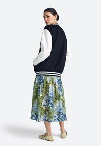 Fekete-fehér varsity kabát bordázott mandzsettákkal, melyet zöld és kék virágmintás szoknyával viselnek. A modell világoszöld cipőt hord.