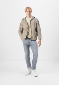 Chaqueta capucha beige clara con cremallera frontal y puños elásticos, combinada con un suéter gris claro, jeans azules y zapatillas blancas.