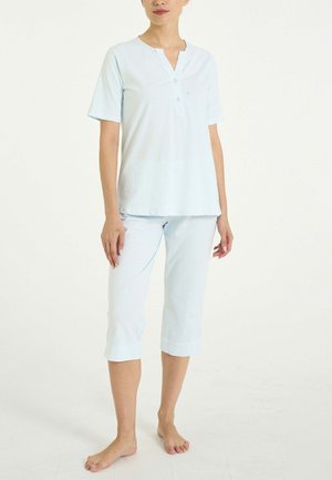 Donna che indossa una maglietta henley a maniche corte azzurro chiaro e pantaloni corti abbinati, in piedi a piedi nudi su uno sfondo bianco uniforme.