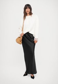 Weiße Bluse mit langen Ärmeln und einer Schleife, schwarze Satin-Maxiröcke und spitze schwarze Ballerinas. Das Model trägt eine gewebte, hellbraune Handtasche.